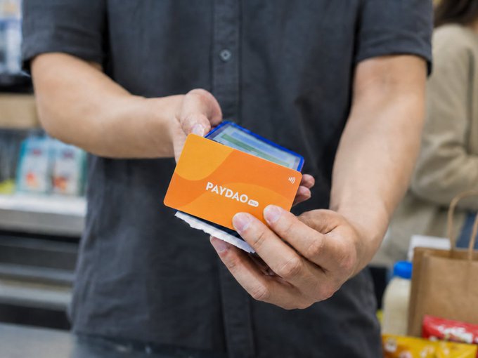 A person holds up an orange "PAYDAO" contactless payment card alongside another card at a checkout counter, visually representing the integration of stablecoin-based payment solutions into ordinary retail transactions. The scene reflects the post's message about embedding digital currency payments seamlessly into everyday commerce, as seen in Lawson's upcoming POS trial in Tokyo.