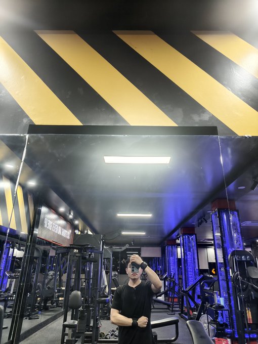 A selfie taken in a gym mirror showing a man taking a photo with his phone, wearing a black polo shirt, with gym equipment and a yellow-striped ceiling visible behind him.