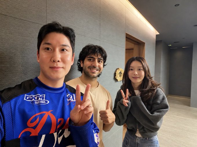 Three people—an East Asian man in a blue 'WILD WING CUSTOM' jersey, a South Asian man in a beige shirt, and an East Asian woman in a gray sweater—pose together making peace signs and smiles.