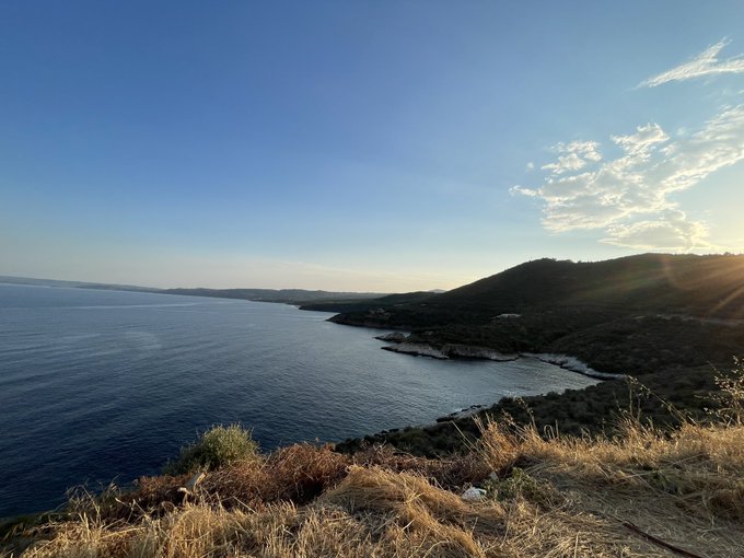 A scenic coastal landscape featuring calm blue waters, distant mountains, and grassy cliffs in the foreground, captured during golden hour with the sun low on the horizon.