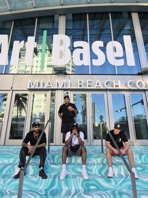 Four people pose on teal-patterned steps in front of a glass building entrance with large "Art Basel" and "MIAMI BEACH CO" signage visible above them.
