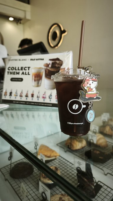 Close-up of an iced coffee drink in a clear plastic cup with a cat keychain charm and brown straw, sitting on a glass counter at what appears to be a Coffee Island café with pastries visible below.