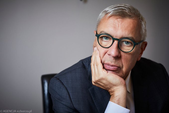 A serious-looking older man with gray hair and teal-framed glasses rests his chin on his hand, wearing a dark checked suit jacket and white shirt. The watermark "© AGENCJA wyborcza.pl" appears in the lower left corner, suggesting the image accompanies a news article about legal proceedings involving Roman Giertych and the ZondaCrypto case mentioned in the post.