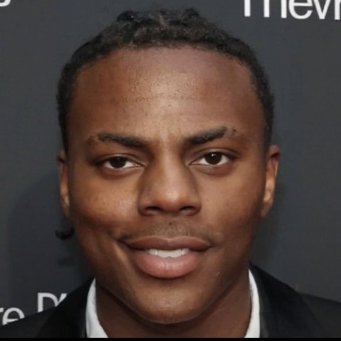 A close-up headshot of a young man with braided hair, wearing a dark suit and white shirt, posing at what appears to be a media event.
