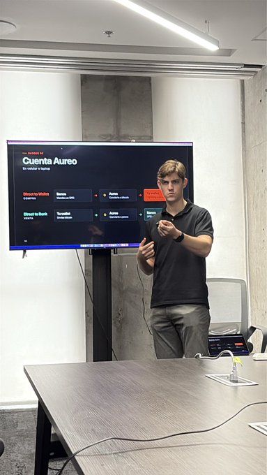 A young man in a dark polo shirt presenting in front of a screen displaying the 'Cuenta Áurea' dashboard with options for 'Cloud to Wallet,' 'Banco,' 'Autos,' 'Bienes,' and 'Cloud to Bank' interactions.