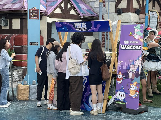 A group of young people gather at a purple promotional booth advertising 'MAGICORE' with Halloween-themed graphics, including witches and characters in costumes, in what appears to be a theme park.