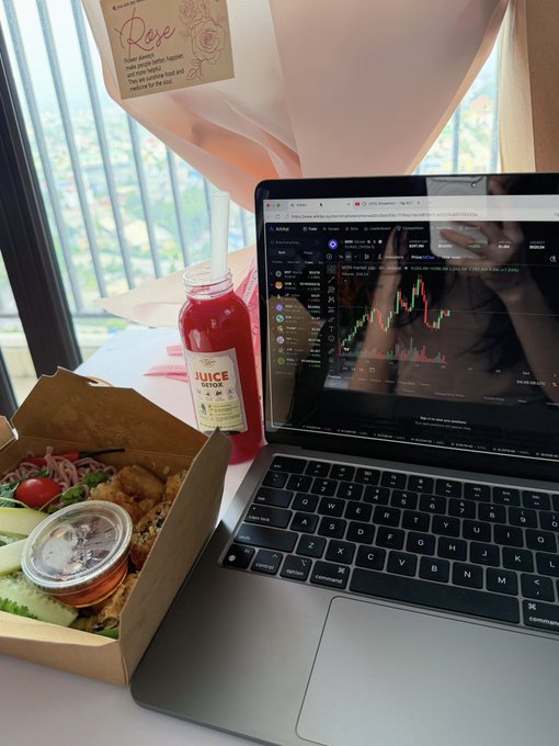 A laptop displaying a trading chart on Coinbase sits next to a healthy meal in a cardboard box and a bottle of juice on a table by a window.