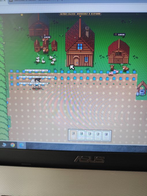 A photograph of a laptop screen displaying a pixel-art farming or village management game with characters, houses, and crop fields.