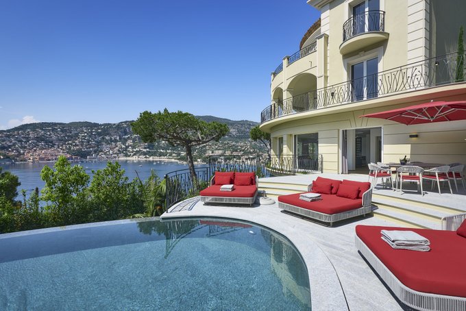A luxury villa terrace overlooking a bay, featuring a swimming pool, red sun loungers with white towels, a red umbrella, and elegant balcony railings.