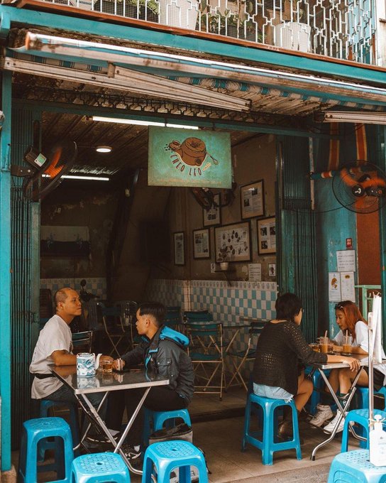 A casual street-side café named 'Cà Phê Quán' with patrons seated on blue plastic stools at small tables under a striped awning.