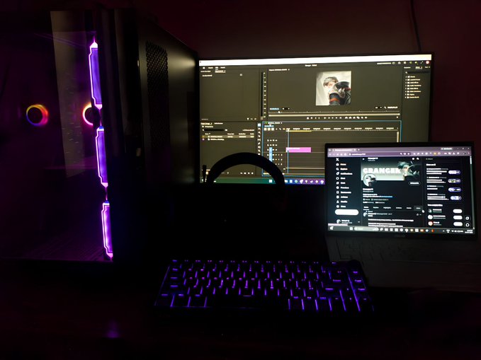 A dimly lit PC gaming/workstation setup featuring a desktop tower with purple LED lighting, a monitor showing video editing software, and a laptop displaying a YouTube channel page.