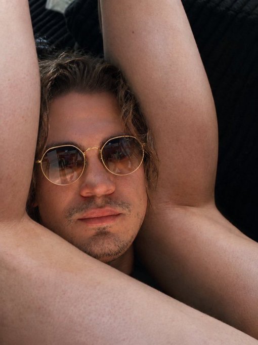 Close-up portrait of a man with curly brown hair, a light beard, and round gold-rimmed sunglasses, with his arms raised behind his head.