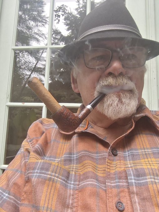 An older man wearing a dark fedora, glasses, and a plaid flannel shirt is smoking a wooden pipe, with visible smoke curling around his face and a white-framed window in the background.