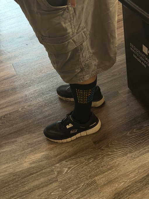 A person stands on a wooden floor wearing olive green cargo shorts, black patterned socks, and black Sketchers sneakers, with a "RESIDENTIAL WASTE SERVICES" bin nearby.