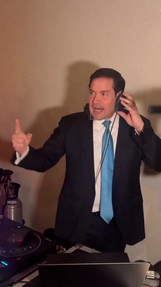 The image features a man wearing a suit, tie, and headset standing behind a laptop computer. He appears to be making a gesture with his hand while holding the headset up to his ear. The background of the photo shows a wall that has a picture hanging on it. The caption "Happy bday rubes" is written in white text at the bottom right corner of the image, which seems to be related to the man's birthday celebration or event taking place around him.
