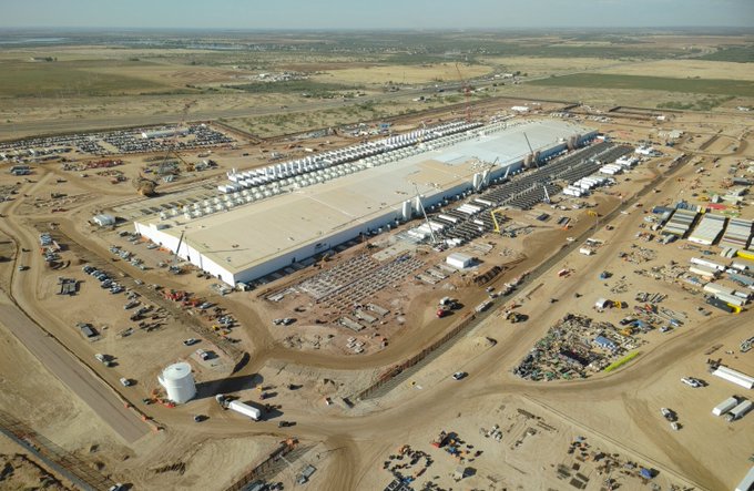 An aerial photograph of a large industrial construction site showing a massive factory building being built in an arid, open landscape with construction equipment and parking areas.
