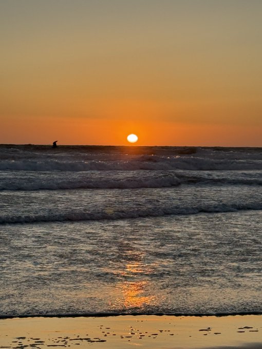 A serene sunset over the ocean, with the sun low on the horizon casting orange reflections on the rolling waves and wet sandy beach in the foreground.