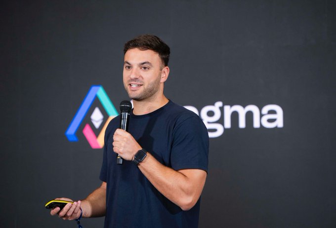 A man in a navy blue t-shirt speaks into a microphone while standing in front of a backdrop displaying the 'Dogma' logo with colorful geometric shapes.