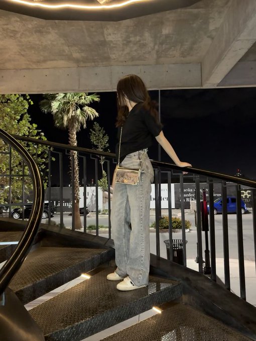 A young woman with long hair stands on outdoor stairs at night, leaning against a metal railing, wearing baggy jeans and a small crossbody bag.