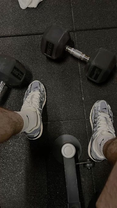 A first-person overhead view of someone at the gym, with white and gray Nike sneakers standing on black rubber flooring near Intek hex dumbbells and a barbell plate.