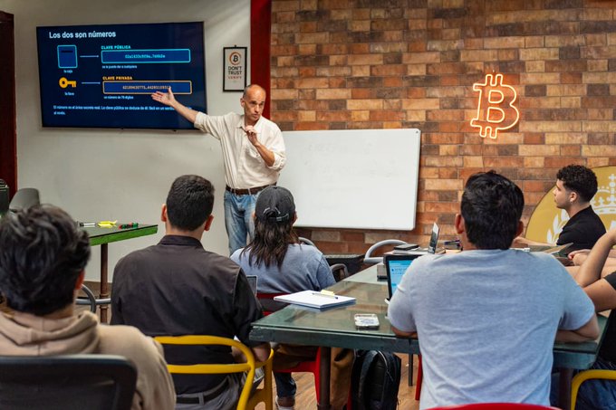 Adam Gibson points to a screen titled "Los dos son números" detailing "CLAVE PRIVADA" and "CLAVE PÚBLICA" sections during a Bitcoin-focused class, where students take notes as he explains the fundamentals of cryptography, including keys, UTXOs, and digital signatures in an accessible, example-driven manner.