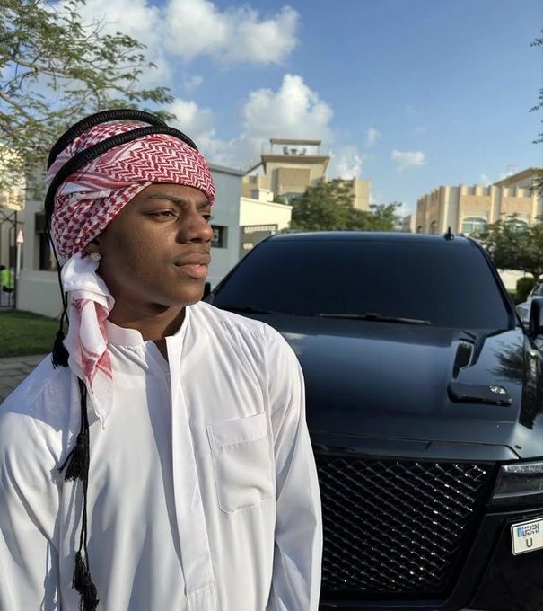 A young man wearing a traditional red and white keffiyeh and white thobe stands in front of a sleek black SUV with a visible "Dubai" license plate, embodying his "up and grinding" weekend hustle in the UAE. The luxury vehicle and Middle Eastern attire reflect both his cultural heritage and successful lifestyle.