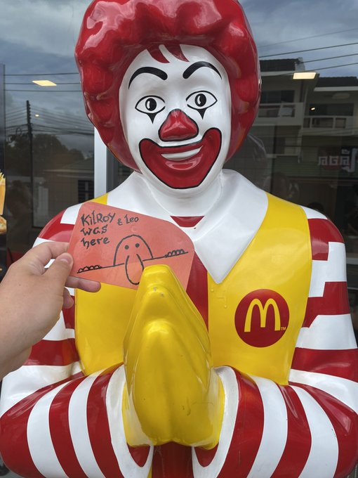 A Ronald McDonald statue stands with hands clasped in prayer as a hand holds up a "Kilroy was here" drawing, blending the nostalgic Kilroy meme with a McDonald's tribute to community members @bliknoiz_06, @mikasasolslayer, @OnlyLJC, @Clive_99, and the $Jotchua, $Kilroy, and $Knight communities.