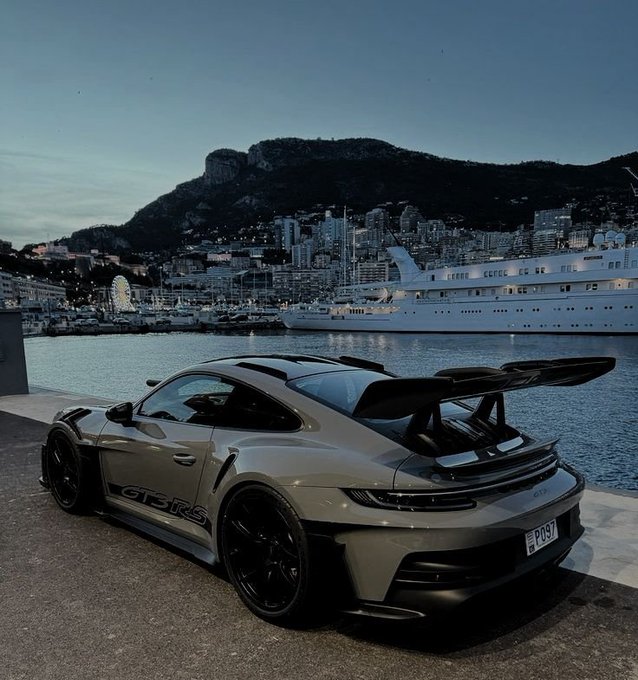 A sleek Porsche 911 GT3 RS with matte black wheels and "GT3 RS" badging is parked on a harbor dock in Monaco, with a large white cruise ship and the city's hillside skyline visible in the twilight background. The luxury sports car exemplifies the post's aspirational message of "dreaming big" beyond budgetary limits, set against one of the world's most exclusive destinations.