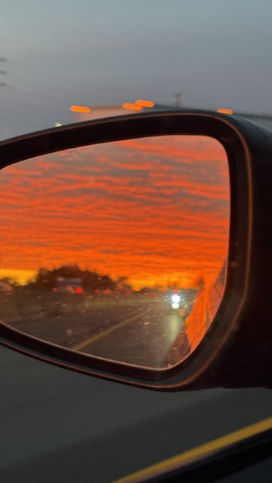 A car's side mirror reflects a vivid orange and red sunset sky, with highway lights visible below.