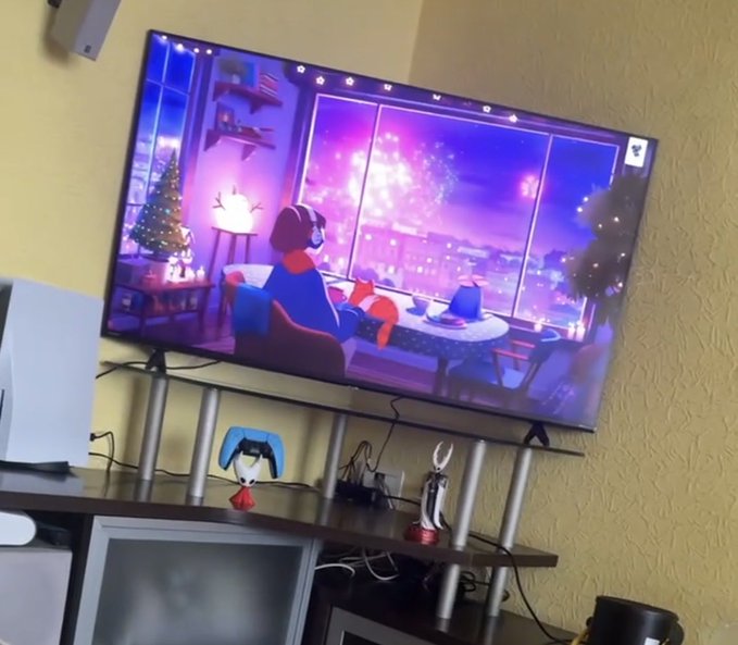 A living room television displays an animated winter scene with a character looking out a window at fireworks and a city, with a PlayStation controller on the entertainment center below.