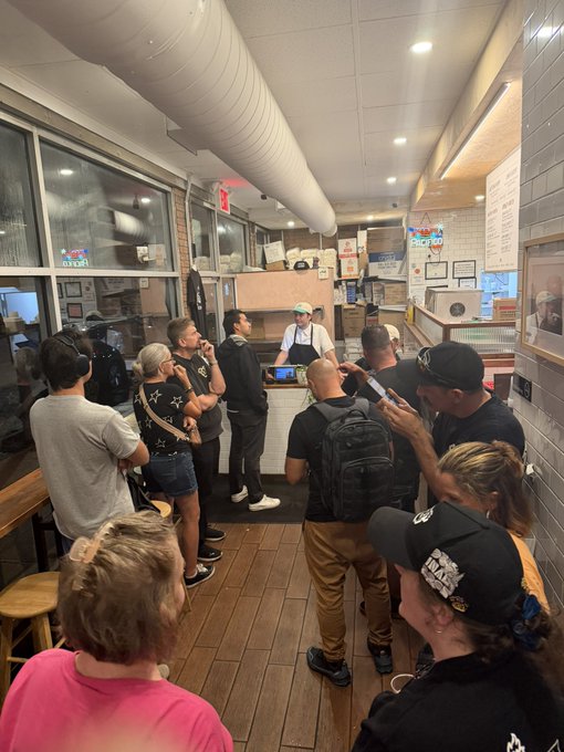 A crowded pizzeria interior with patrons lined up at the counter where a worker in an apron and cap stands, surrounded by exposed ductwork and menu boards.