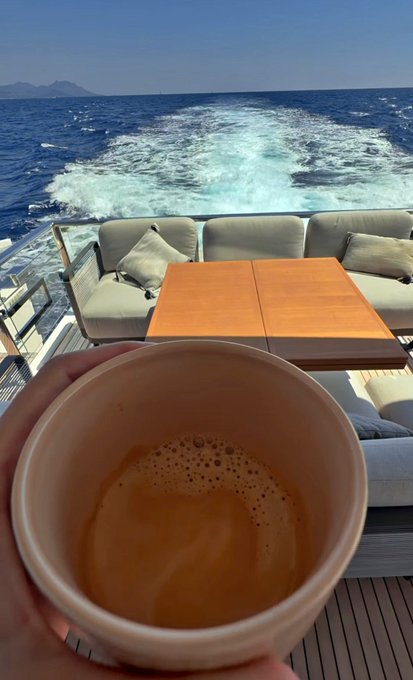 A hand holds a paper cup of coffee overlooking the wake of a boat, with an outdoor seating area featuring beige cushions and wooden tables in the background.