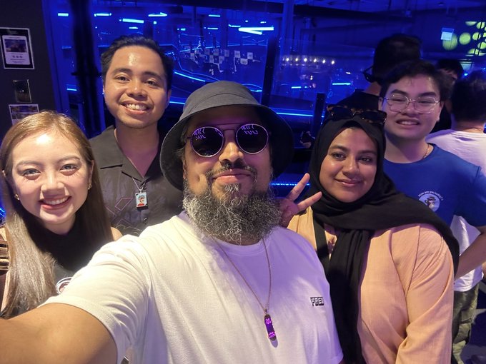 A group selfie taken inside a venue with blue lighting shows five smiling people, including a man in a bucket hat and sunglasses in the center.