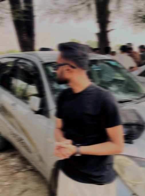 A blurry, motion-effected photo of a man with dark hair and sunglasses wearing a black t-shirt, standing next to a white car with other people visible in the background.