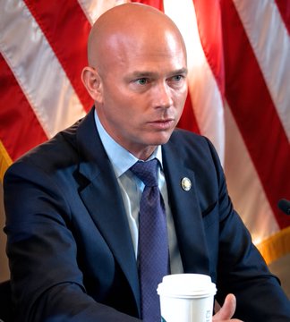 U.S. Representative William Timmons, dressed in a dark suit and blue tie, sits in front of an American flag, appearing focused during a congressional session. The image aligns with his statements advocating for the passage of the Clarity Act to maintain the U.S. economy's global leadership.