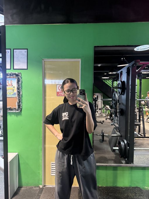 A woman wearing glasses and a black "Chog" t-shirt poses for a mirror selfie in a gym with bright green walls and weight racks visible behind her, promoting the ChogNFT mint for just 10k $chog at the contract address 0x350035555E10d9AfAF1566AaebfCeD5BA6C27777.