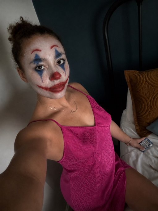A woman wearing clown-style face makeup with red lips and a red nose, dressed in a pink slip dress, takes a selfie in a bedroom setting.