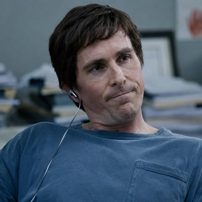 Christian Bale, wearing a blue t-shirt and an earpiece with a white cord, is shown in what appears to be a scene from a movie, looking to the side with a subtle smirk.