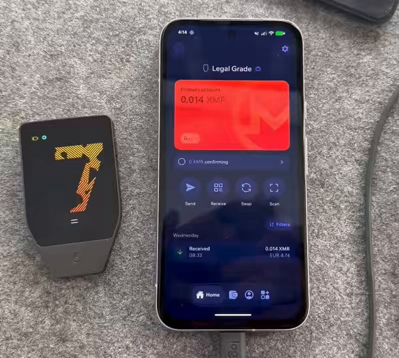 The image shows a smartphone with an app called "Monero" displayed on its screen, along with a wallet icon next to it. The phone appears to be connected to the wallet via Bluetooth technology. The background of the image features a gray surface, which could be a table or countertop. There is also a small amount of text visible in the image that reads: "Legal Grade".