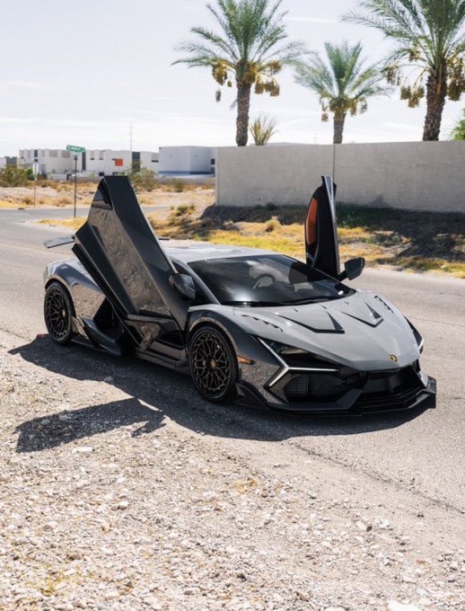 A dark gray Lamborghini Revuelto with its scissor doors open parked on gravel near palm trees.