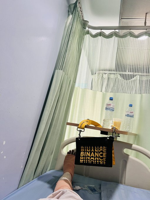 A patient lies in a hospital bed with a leg bandage, their foot resting near a black tote bag branded with "BINANCE BINANCE BINANCE" in yellow text, positioned on the bed rail alongside two water bottles behind a privacy curtain. The image illustrates the touching moment when the patient reunited with their old Binance merchandise, now repurposed by their mother as a small wallet for everyday use.