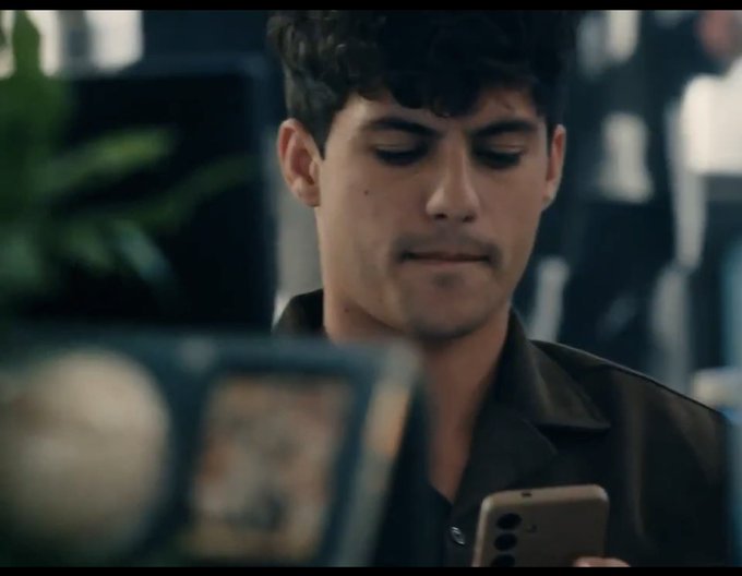 A young man with dark curly hair, identified as "Ron," looks down at his phone with a focused expression, wearing a dark jacket. The image appears to be a still from the PolyMarket commercial mentioned in the post, showing him engaging with the platform while LeBron James is referenced in the same advertisement.