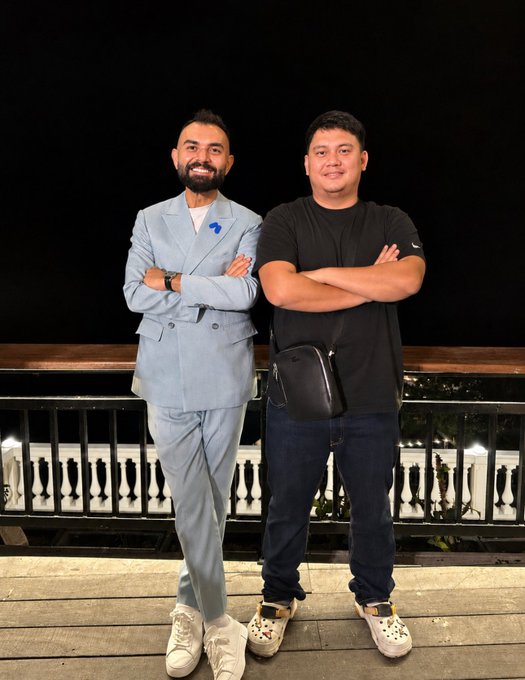 Two men stand with crossed arms on an outdoor balcony at night; one wears a light blue suit and the other wears a black t-shirt with a crossbody bag.