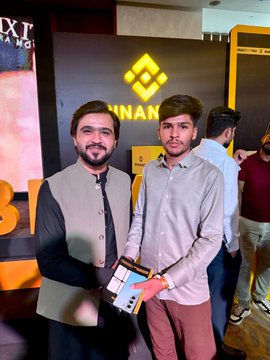 Two young men posing together at what appears to be a Binance event, with one holding an award or plaque, standing in front of a Binance logo backdrop.