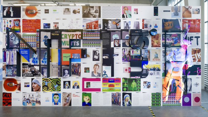 A large colorful wall display or collage featuring the words 'NFT', 'CRYPTO MUTTS', and 'PUSSYC', composed of numerous small images, text blocks, and artistic elements in a gallery setting.