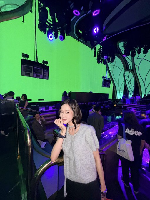A photograph of a young woman with shoulder-length dark hair posing in what appears to be a theme park queue or ride area, with a bright green screen/background, purple stage lights overhead, and other visitors visible behind her near signage for 'Kingdom Callo'.
