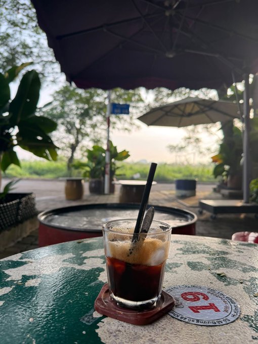 A glass of iced coffee with a spoon on a weathered green outdoor table, set under a large umbrella with a rural landscape in the background.