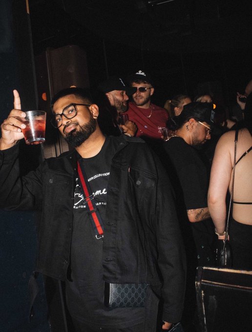 A bearded man in a black jacket and glasses holds up a drink while posing at a crowded, dimly lit nightclub or party venue.