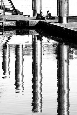 A black-and-white photograph captures two people sitting on a stone platform beside a reflecting pool, with the columns and architectural elements mirrored in the rippling water below. The image's serene, contemplative mood of quiet companionship pairs well with the artwork's title, "We Are Friends."