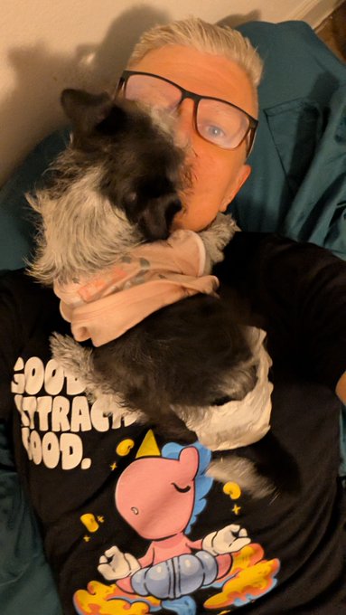 A selfie of a person wearing glasses and a dark t-shirt with a cartoon character, cuddling a black and white dog under a hooded blanket.
